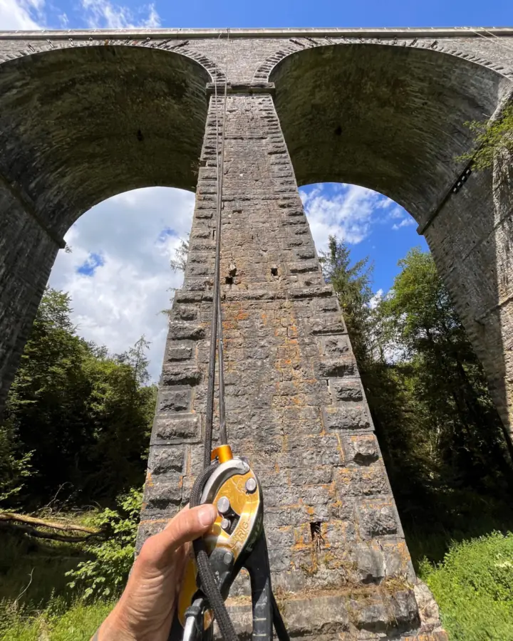 Main d'un cordiste sur un descendeur, corde engagée, au pied d'un viaduc en pierre